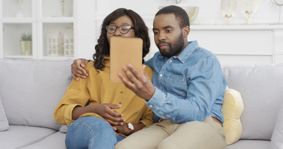 Young African American couple sitting on couch in living room and videochatting on tablet device. Male and female talking via webcam on computer at home on white sofa. Man and woman having videochat. Royalty-Free Stock Footage #1055589971