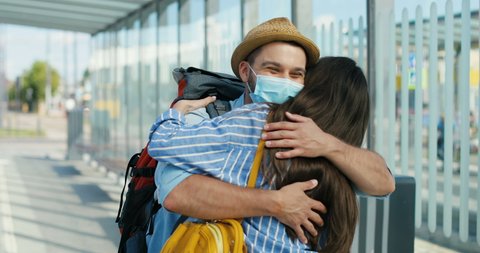 Joyful Caucasian young couple in medical masks meeting at train station and hugging on summer day. Happy handsome man meet beautiful woman at bus stop. Coming back from trip. Coronavirus pandemic.
