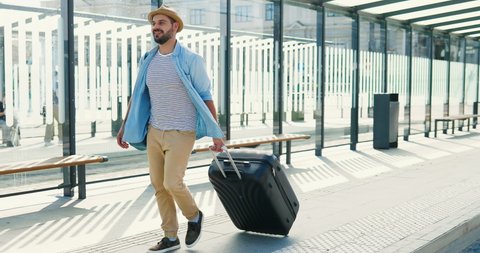 Caucasian young stylish man traveller in hat and with backpack walking at bus station and carrying suitcase on wheels. Handsome male tourist strolling outdoor from train station or aeroport.