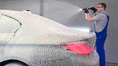 Car with turned on backlights standing in car wash service. Worker cover the car with foam with high-pressure sprayer.