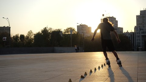 Young long-haired man roller skater is dancing between cones in evening city square at sunset. Freestyle slalom Roller skating boy in slow motion. Stylish urban guy spends free leisure time in sport