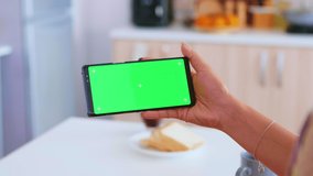 Woman watching a video on phone with green screen in kitchen during morning. Green screen chroma mock up isolated mockup background ready to be replaced with your text, logo or advertisement. Using