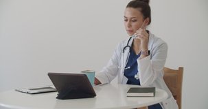 Lunch break in a medical facility. Side view of cute young doctor in white coat working at tablet answering an incoming call, on table next to cup with hot drink. 4k raw video footage slow motion