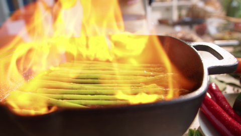 Close-up shot of professional chef igniting the fire with lighter on grill pan and then using metal tongs for flipping asparagus during flambeing