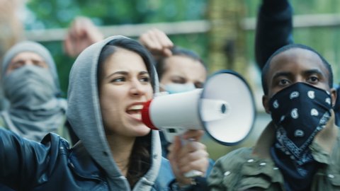 Caucasian young woman leader screaming mottos in megaphone at street in crowd and smoke at protest against racism in USA. Active mixed-races protesters shouting demands. Female leading at rebellion.