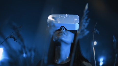Young woman uses virtual or augmented reality glasses in magic atmosphere of blue neon rays, female VR headset user at digital interactive art performance, entertainment of future