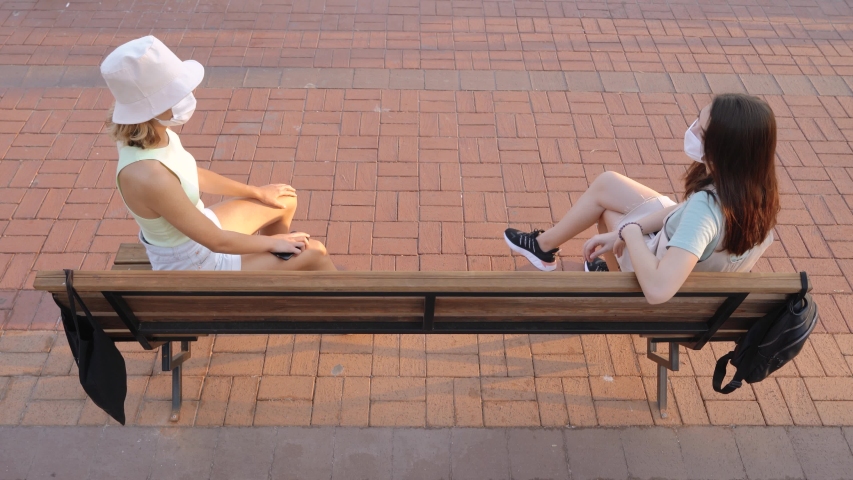 Attractive young women in social safe distance sitting on bench and speaking.
