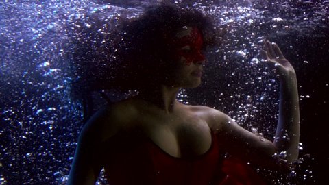 Close-up portrait of a beautiful girl who is under water, she has a lush chest and a red dress fluttering with fabrics. It floats on a dark background with bubbles around it.