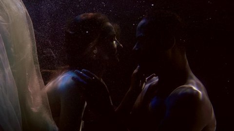 Close-up portrait of a tender couple in love who are under water and swim beautifully, a girl in a dress with a fluttering fabric and a dark-skinned man. They are on a dark background with bubbles.