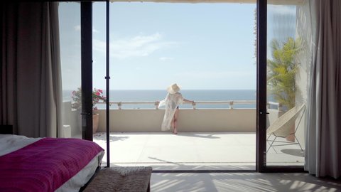 Woman in bikini and sheer dress stands on her balcony as seen from her bedroom taking in the beautiful view and sunshine while on vacation