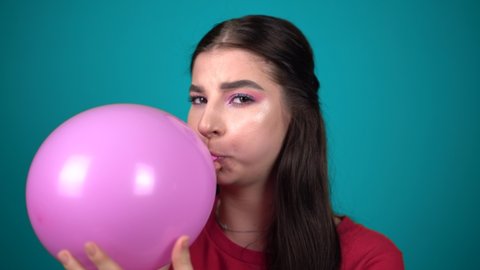 Young woman inflates a balloon isolated on blue background Young happy woman looking at camera and inflates a pink balloon clos up portrait