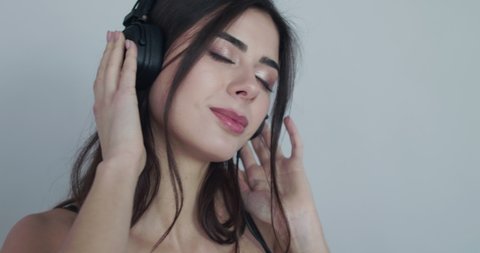 Portrait of nude girl in bra listening to the music with closed eyes and swaying. She hanging headphones around her neck, looking into and biting a lip at camera on background. Slowly in 4K