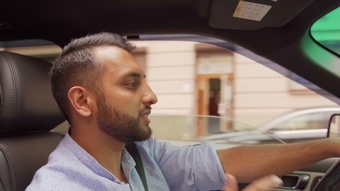 Tilt up side view of young Middle Eastern man rapping to music while driving car down city road with open window