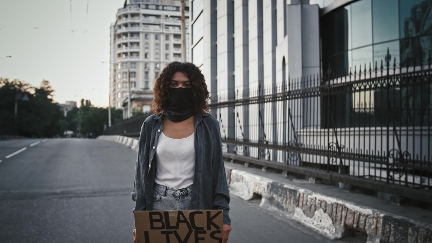 Dark-skinned girl in black bandana on face. Showing cardboard tablet with inscription Black lives matter, standing on bridge. Protests in USA