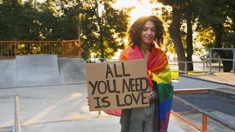 Afro-american female wrapped in LGBT pride flag. Showing cardboard poster, inscription all you need is love. Posing at sunny skatepark. Slow motion