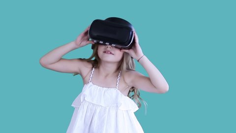 Cute beautiful kid in white dress wearing VR glasses, moving energetically, putting off this helmet, looking scared at camera. Concept of lifestyle. Isolated on blue background Video de stock