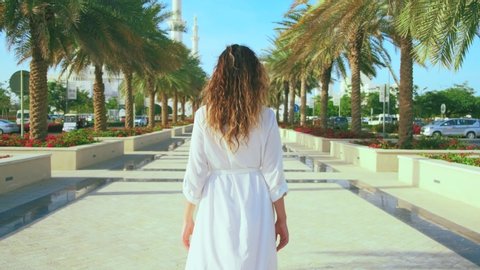 Beautiful cityscape tourism city Abu Dhabi. Silhouette Young happy woman tourist turned away, walk town alley, enjoy Arabian sights. Long white strict dress sleeves. Dubai UAE 4k. Girl back rear view