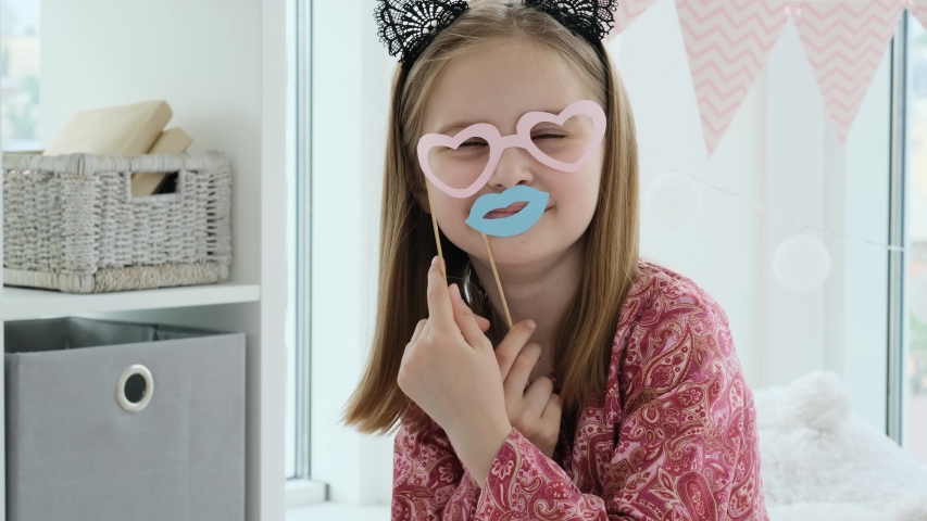 Little girl playing with paper glasses and lips in a kids room