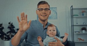 happy father waving hand while holding infant son and having video call