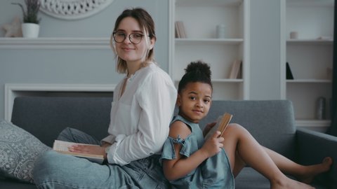 Portrait of happy beautiful family young adult single mother foster parent and african daughter. Sit on couch back to back in living room of house. With book in their hands smiling looking at camera.の動画素材