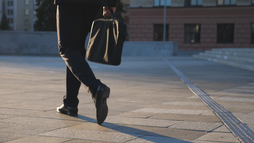 Feet of young businessman with a briefcase walking in city street. Business man commuting to work. Confident guy in suit being on his way to work. Cityscape background. Slow motion Rear view Close up Royalty-Free Stock Footage #1055675858