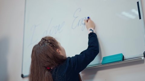 KYIV, UKRAINE - May 2020: Schoolgirl writes on a board. Back view of a little girl in a school uniform is writing on a whiteboard with a marker. Primary education concept.
