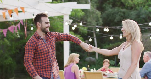Caucasian couple spending time in garden together, dancing and smiling, with their family sitting at dinner table in the background, in slow motion.