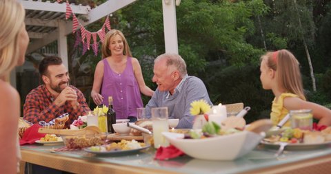 Multi-generation Caucasian family spending time in garden together, sitting at a dinner table, the senior woman kissing her husband, in slow motion.