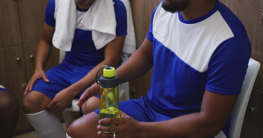 Two mixed race male soccer players in a dressing room talking and holding bottle of water in slow motion. Sports team taking a break from a match.