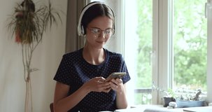 Student teen girl wears headphones and eyeglasses sit indoors use cell phone text message to friend, chatting on-line discuss news with classmate, enjoy distant communication in social network concept