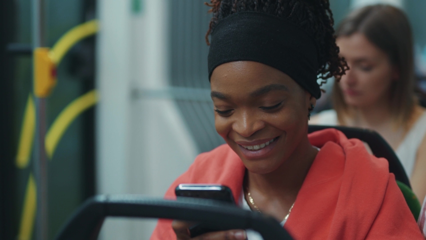 Charming afro-american woman with smartphone read messages from boyfriend smiling in the tram. Public transport. | Shutterstock HD Video #1055713640