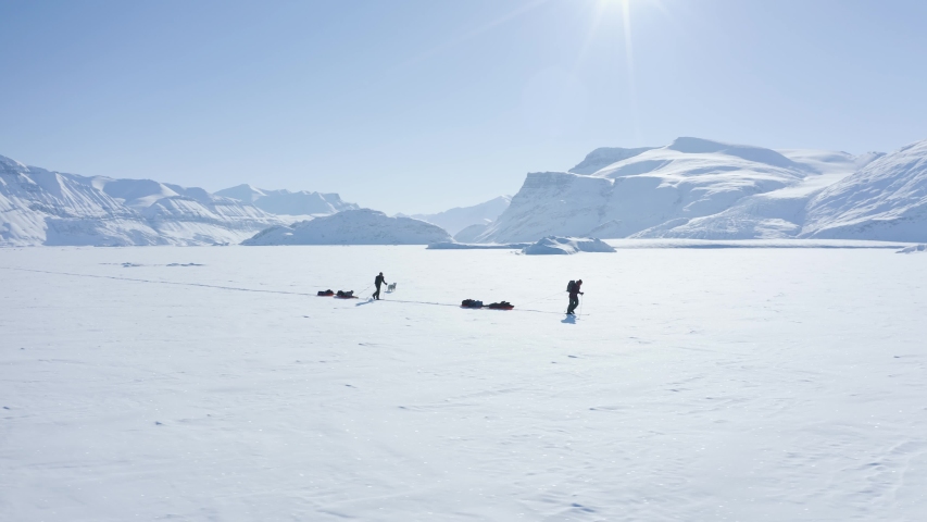 Two extreme sports athletes and a husky dog are hiking in the middle of the arctic on a frozen fiord surrounded by snowy, white and cold mountain peaks towards north pole, pulling bulka sledges | Shutterstock HD Video #1055713874