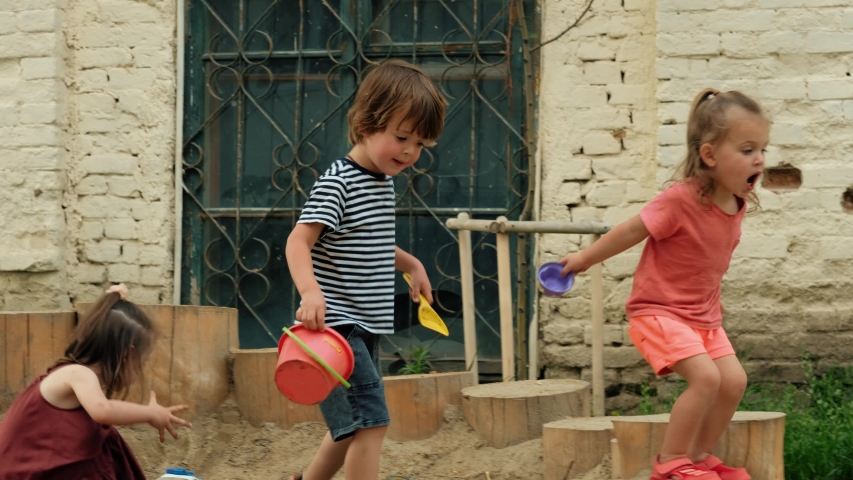 Side view of active little children in casual clothes with plastic toys jumping on wooden logs while playing together in sandy playground in summer day