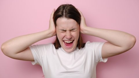 Irritated young woman frustrated loud noise covering ears clasping head, want to stop unpleasant voices in mind. Girl angry with her neighbours who are very noisy, isolated on pink studio background