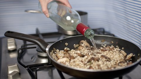 Pouring white dry wine into the mushroom risotto. Concept of preparing and adding an ingredient into a mushroom risotto on a pan on the gas stove.