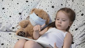 A small child with a bear in a mask plays a tablet while lying on the bed. Kovid-19. Kid is quarantined at home. Distance learning for preschoolers. Modern technologies.