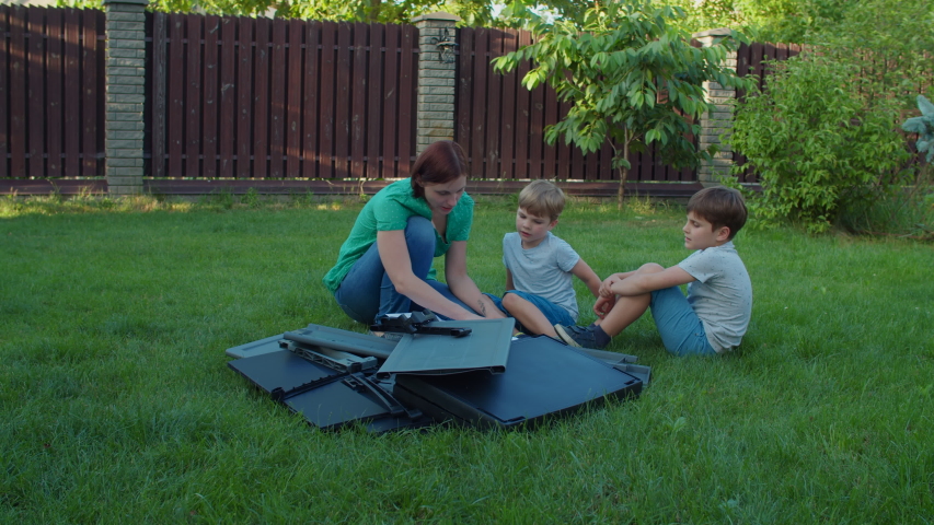 Family of young single mother and two kids assembles furniture with instruction in backyard on sunny day. Happy family weekend activities outdoors. Slow motion, steadicam shot.