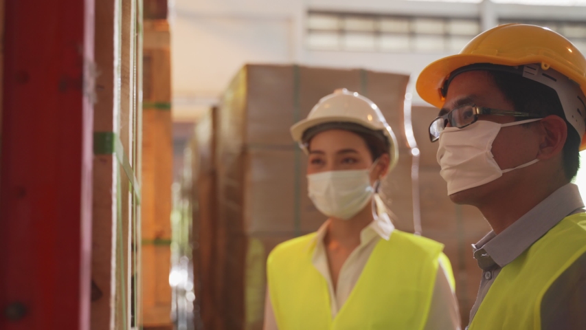 Asian female worker talking with manager in warehouse store. Woman and man engineers people wear safety hard helmet, vest and face mask checking storage box parcel in factory during covid 19 pandemic. Royalty-Free Stock Footage #1055786942
