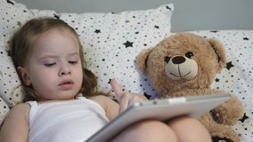 Little happy child plays a tablet while lying on a bed with a teddy bear. The kid watches the children's channel through the touch monitor. Teaching a preschooler remotely online. Close-up.