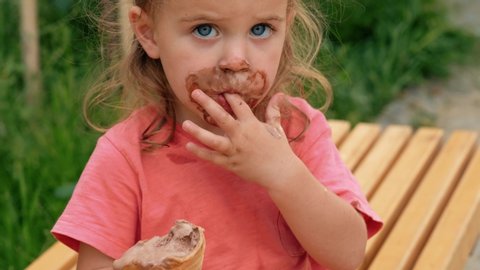 Cute little girl in casual clothes with face smeared with chocolate ice cream licking fingers while sitting on bench in park in summer day