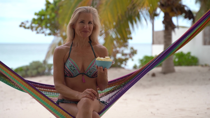 Pretty mature woman in sunglasses and bikini in a hammock eating pineapple and looking like it is unbelievably appetizing.