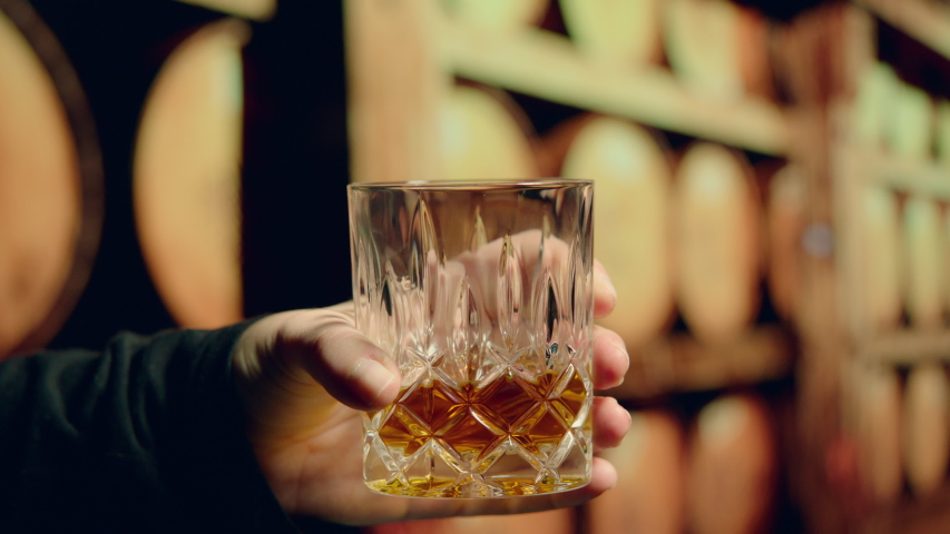 Looking Carefully at Bourbon- Slow Motion Hand Swirling Glass of Bourbon Whiskey inside of Still House with Whiskey Barrels in Background