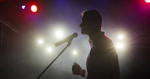 Silhouette of man stand up comedian telling funny jokes in micropphone standing on stage. Successful performance in spotlights on dark background, side view. Comedian at his concert.