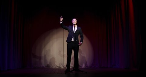 Man in suit stand up comedian speaking jokes in micropphone standing on stage. Funny performance. Comedian clapping hands overhead and calling audience doing the same.