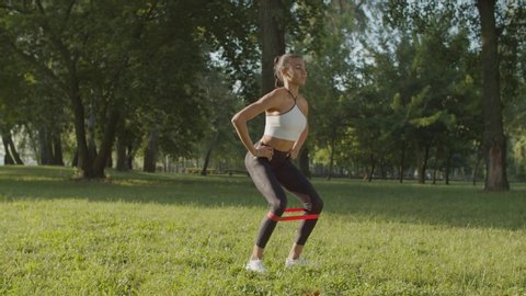 Beautiful sporty fitness african american woman squatting doing sit-ups with resistance band in public park at sunrise. Active motivated female athlete working out outdoors, crouching using loop band.