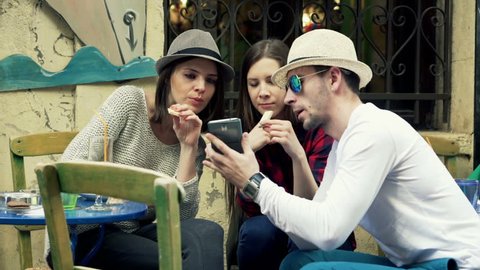 Young friends talking and using smartphone sitting in cafe in city 
