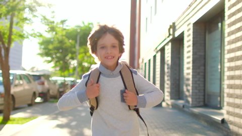 Portrait of happy school boy smiling schoolboy little boy child with backpack alone going running to children kid at sun street outdoors, First-grade student primary first class education 4 K slow-mo