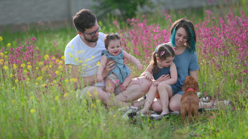 A happy young family is relaxing on a flowering field with his beloved dog. Father and mother play with children in a field on a summer sunny day. Family having fun. Royalty-Free Stock Footage #1055807522