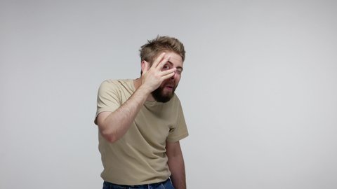 Funny curious nosy bearded guy in t-shirt looking through fingers with prying eyes, spying, cautiously watching forbidden secret, peeking scared shy. indoor studio shot isolated on gray background