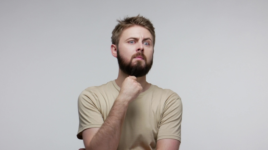 Pensive confused guy leaning hand, rubbing beard while thinking over difficult choice, imagining plan in mind, having doubts, pondering idea looking puzzled clueless. indoor studio shot isolated | Shutterstock HD Video #1055808383